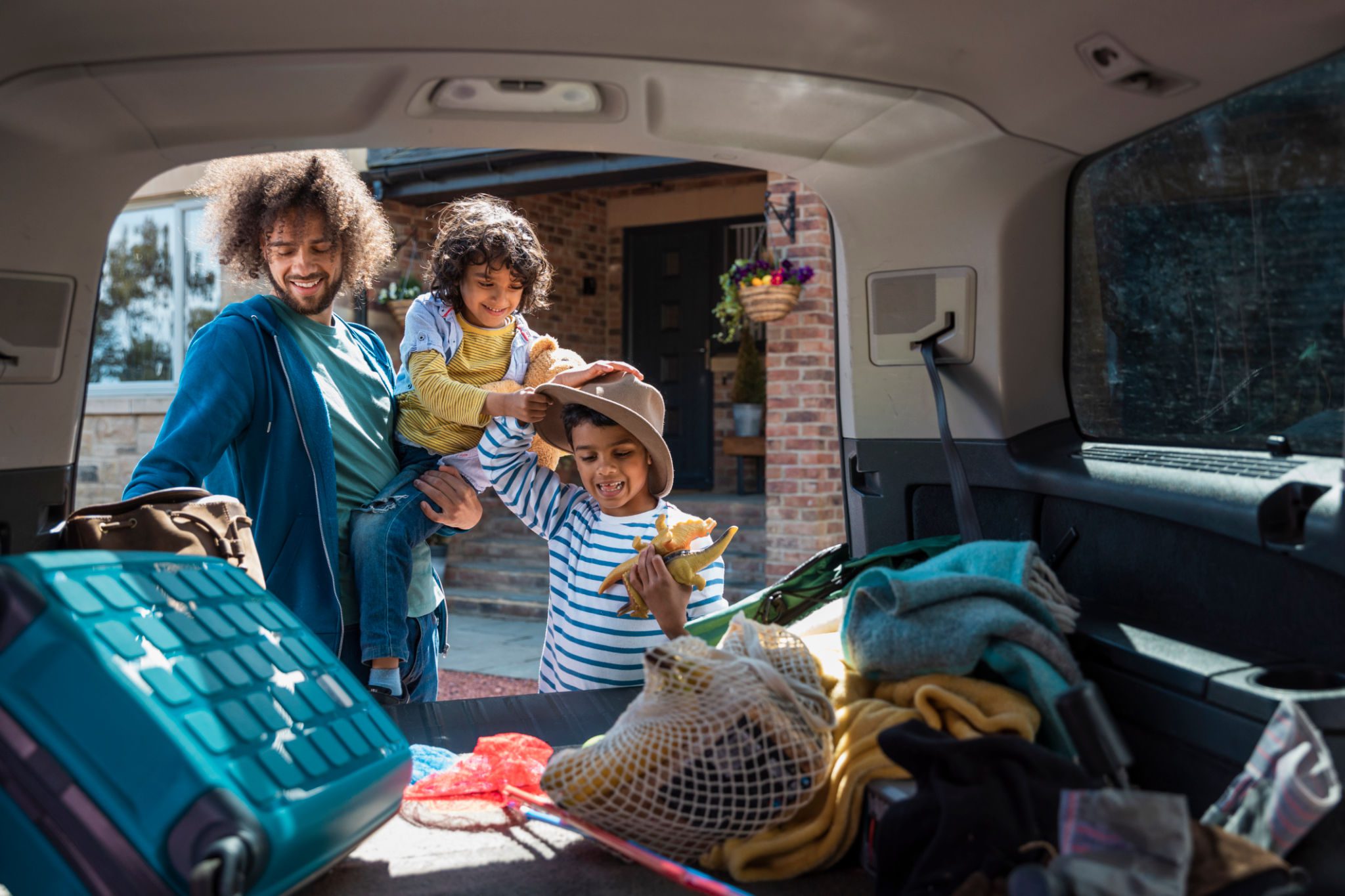 Medium shot with camera placed inside of a car showing a man and his son unpacking a car boot, getting their bags and toys when they arrive at the lodge they're staying on during a staycation in the Northeast of England together. The young boy is trying on his fathers hat, having fun and the man is holding one of the boys.